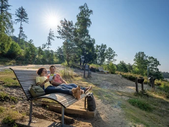 Oerlinghausen-Sandgrube-Hassler-Teutoburger-Wald-Tourismus-Patrick-Gawandtka-011_lowres.jpg Zwei Personen entspannen auf einer Holzbank in einer sonnigen Waldlichtung im Teutoburger Wald.