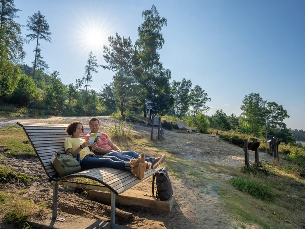 Oerlinghausen-Sandgrube-Hassler-Teutoburger-Wald-Tourismus-Patrick-Gawandtka-011_lowres.jpg Zwei Personen entspannen auf einer Holzbank in einer sonnigen Waldlichtung im Teutoburger Wald.
