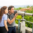 Eine Frau und ein Mann betrachten durch ein Fernrohr in einem grünen Waldgebiet eine Landschaft.