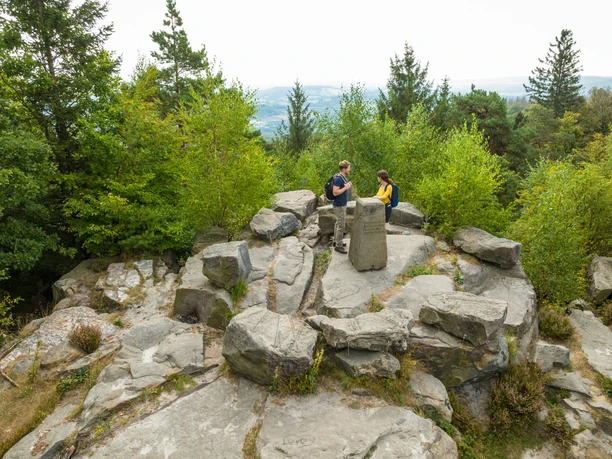 Zwei Wanderer bei einer Pause auf einem Felsenplateau mit Aussicht inmitten eines bewaldeten Gebiets.