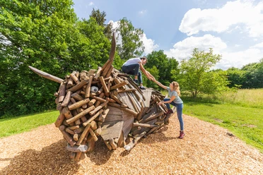 Warburg-Hammerhof-Teutoburger-Wald-Tourismus-A-Röser-018.jpg Kinder klettern auf einer kreativen Holzskulptur in einer sonnigen, grünen Landschaft im Wald.