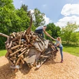 Warburg-Hammerhof-Teutoburger-Wald-Tourismus-A-Röser-018.jpg Kinder klettern auf einer kreativen Holzskulptur in einer sonnigen, grünen Landschaft im Wald.