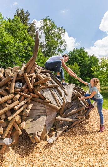 Warburg-Hammerhof-Teutoburger-Wald-Tourismus-A-Röser-018.jpg Kinder klettern auf einer kreativen Holzskulptur in einer sonnigen, grünen Landschaft im Wald.
