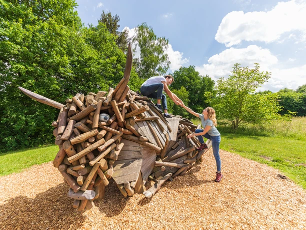 Warburg-Hammerhof-Teutoburger-Wald-Tourismus-A-Röser-018.jpg Kinder klettern auf einer kreativen Holzskulptur in einer sonnigen, grünen Landschaft im Wald.
