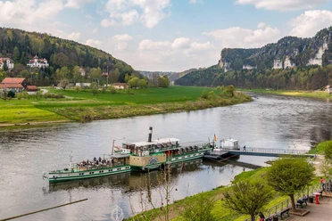 Een groene raderstoomboot ligt aangemeerd aan een steiger op een rivier, omringd door groene heuvels en beboste rotsen onder een bewolkte hemel.