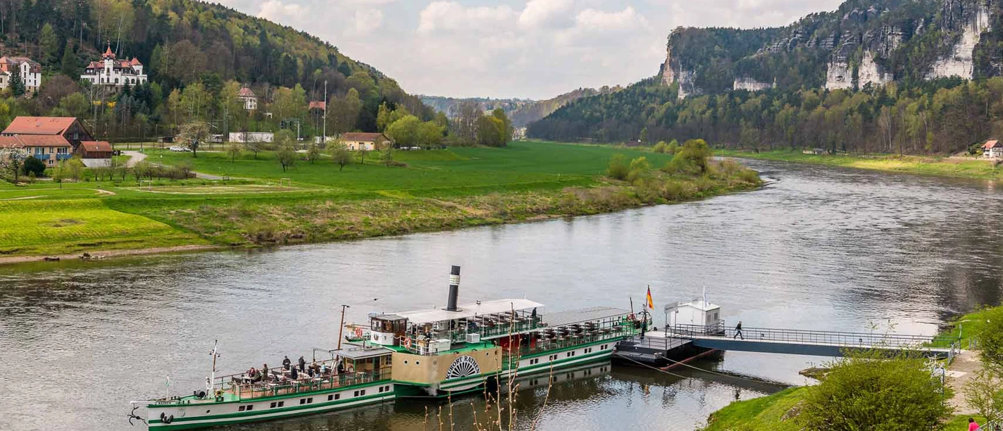 A green paddle steamer is moored on a jetty on a river, surrounded by green hills and wooded rocks under a cloudy sky.
