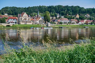Anlegestelle Sächsische Dampfschifffahrt Wehlen Flusslandschaft mit kleinen Booten am Ufer, im Hintergrund eine malerische Stadt mit Fachwerkhäusern und einer Kirche, umgeben von grünen Hügeln.