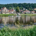 Anlegestelle Sächsische Dampfschifffahrt Wehlen Flusslandschaft mit kleinen Booten am Ufer, im Hintergrund eine malerische Stadt mit Fachwerkhäusern und einer Kirche, umgeben von grünen Hügeln.