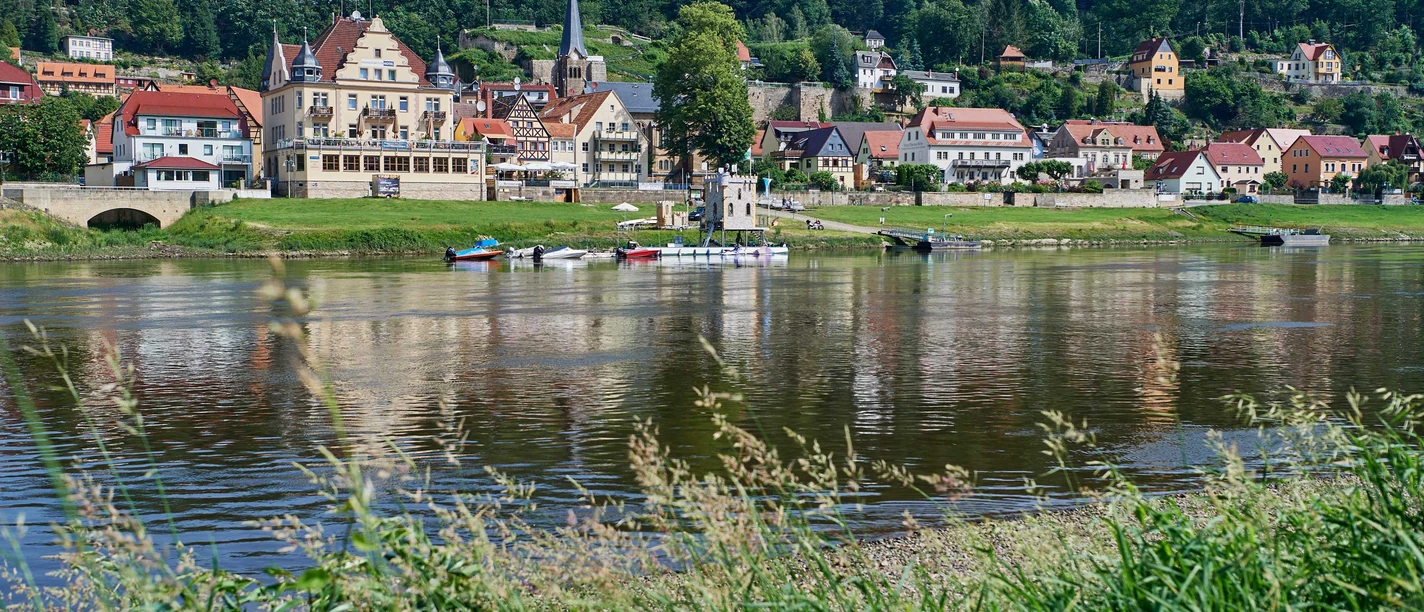 Anlegestelle Sächsische Dampfschifffahrt Wehlen Flusslandschaft mit kleinen Booten am Ufer, im Hintergrund eine malerische Stadt mit Fachwerkhäusern und einer Kirche, umgeben von grünen Hügeln.