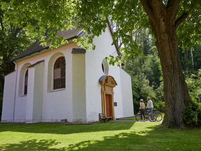 Zwei Radfahrer betrachten eine kleine, weiße Kapelle im Grünen, umgeben von Bäumen.