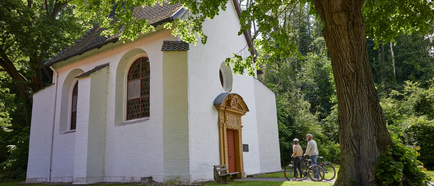 Borchen-Kluskapelle-Teutoburger-Wald-Tourismus-T-Evers-110.jpg Zwei Radfahrer betrachten eine kleine, weiße Kapelle im Grünen, umgeben von Bäumen.