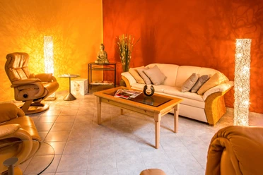 Gezellige woonkamer met beige bank, bruine fauteuil, glazen tafel en Boeddhabeeld. Warme verlichting en oranje gekleurde muren creëren een ontspannen sfeer.