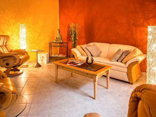 Wohlfühl-Vitallounge im Parkhotel Gemütliches Wohnzimmer mit beigem Sofa, braunem Sessel, Glastisch und Buddha-Statue. Warme Beleuchtung und orangefarbene Wände schaffen eine entspannte Atmosphäre.