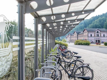 Fahrradständer am Nationalparkbahnhof Bad Schandau Überdachter Fahrradständer mit mehreren Fahrrädern, daneben ein Gebäude mit rosa Fassade und grüner bewaldeter Hintergrund.Covered bicycle stand with several bicycles, next to it a building with a pink facade and a green wooded background.Krytý stojan na kola s několika koly, vedle něj budova s růžovou fasádou a zeleným zalesněným pozadím.Zadaszony stojak rowerowy z kilkoma rowerami, obok budynek z różową fasadą i zielonym zalesionym tłem.Overdekte fietsenstalling met verschillende fietsen, daarnaast een gebouw met een roze gevel en een groene beboste achtergrond.Bancarella coperta con diverse biciclette, accanto a un edificio con la facciata rosa e uno sfondo verde e boscoso.