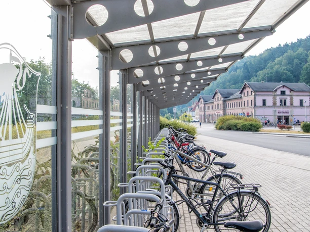 Fahrradständer am Nationalparkbahnhof Bad Schandau Überdachter Fahrradständer mit mehreren Fahrrädern, daneben ein Gebäude mit rosa Fassade und grüner bewaldeter Hintergrund.