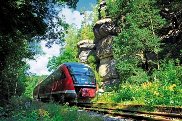 Een rode trein rijdt door een groen, bebost landschap met grote rotsen op de achtergrond onder een blauwe lucht.