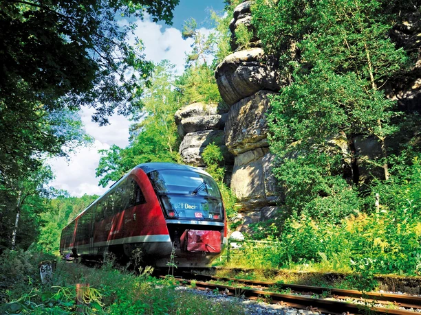 Een rode trein rijdt door een groen, bebost landschap met grote rotsen op de achtergrond onder een blauwe lucht.