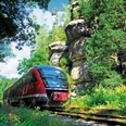 Nationalparkbahn U28 Ein roter Zug fährt durch eine grüne, bewaldete Landschaft mit großen Felsen im Hintergrund unter einem blauen Himmel.A red train travels through a green, wooded landscape with large rocks in the background under a blue sky.Červený vlak projíždí zelenou zalesněnou krajinou s velkými skalami v pozadí pod modrou oblohou.Czerwony pociąg jedzie przez zielony, zalesiony krajobraz z dużymi skałami w tle pod błękitnym niebem.Een rode trein rijdt door een groen, bebost landschap met grote rotsen op de achtergrond onder een blauwe lucht.Un treno rosso attraversa un paesaggio verde e boscoso con grandi rocce sullo sfondo, sotto un cielo blu.