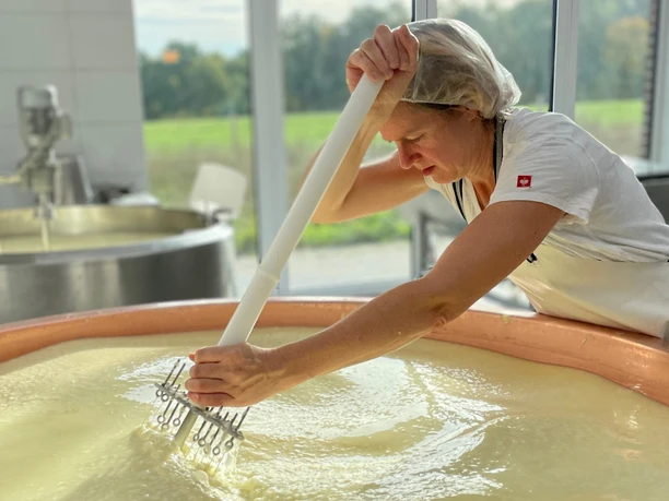 Eine Käserin in weißer Arbeitskleidung rührt konzentriert in einem großen Kupferkessel mit Milch im Sonnenlicht der Fenster im Hintergrund.