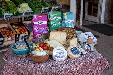 Bauckhof - Sortiment Das Bild zeigt eine liebevoll arrangierte Auswahl an biologischen Backwaren, verschiedenen Käsesorten und Müsli-Schalen, die appetitlich auf einem Tisch drapiert sind. Im Hintergrund sind frisches Bio-Gemüse und Obst zu sehen, die die Vielfalt und Frische des Sortiments unterstreichen.