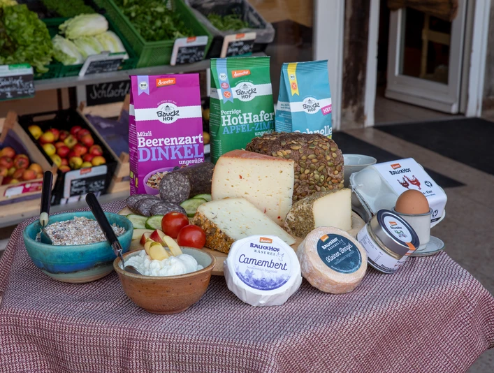 Bauckhof - Sortiment.jpg Das Bild zeigt eine liebevoll arrangierte Auswahl an biologischen Backwaren, verschiedenen Käsesorten und Müsli-Schalen, die appetitlich auf einem Tisch drapiert sind. Im Hintergrund sind frisches Bio-Gemüse und Obst zu sehen, die die Vielfalt und Frische des Sortiments unterstreichen.