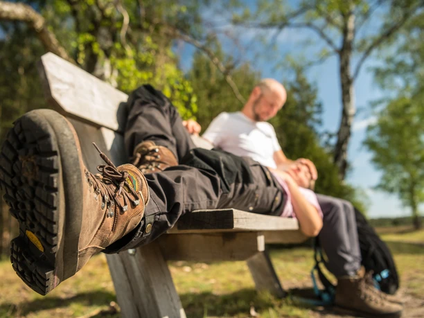 Wandererpaar ruht auf einer Holzbank im Wald, Fokus auf einem Wanderschuh im Vordergrund.