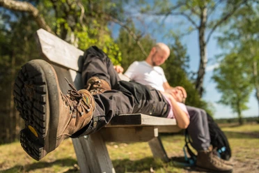 Enstpannung auf dem Königinnenweg Wandererpaar ruht auf einer Holzbank im Wald, Fokus auf einem Wanderschuh im Vordergrund.
