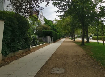 Ein ruhiger Gehweg, flankiert von Bäumen und Grün, neben einer gepflegten Wohnanlage.