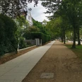 Ein ruhiger Gehweg, flankiert von Bäumen und Grün, neben einer gepflegten Wohnanlage.