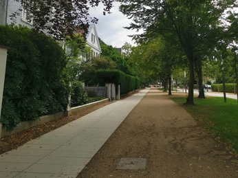 ©Mechthild Klein.jpg Ein ruhiger Gehweg, flankiert von Bäumen und Grün, neben einer gepflegten Wohnanlage.