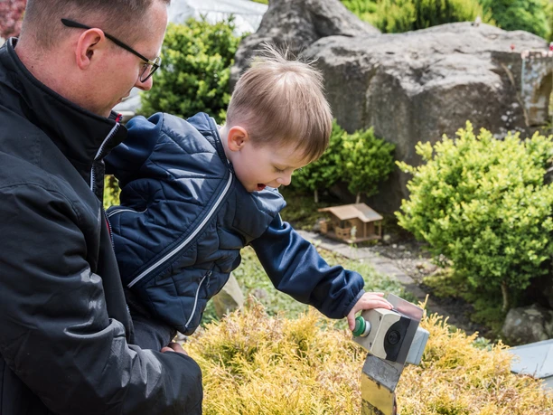 A man lifts up a child in a blue jacket who joyfully presses a green button in a garden with rocks and bushes.