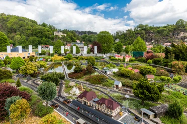 Miniature landscape with model railroad, surrounded by colorful buildings and green trees, under a cloudy sky.