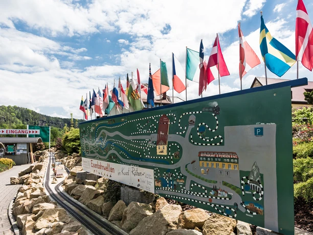 Eisenbahnwelten im Kurort Rathen Wegweiser mit bunten Flaggen und einer Karte, die eine Miniatureisenbahnstrecke zeigt, daneben ein Eingangsschild; sonniger Tag mit blauem Himmel.