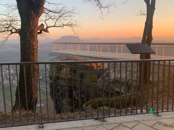 Bastei Aussicht zum Sonnenuntergang Aussichtsplattform bei Sonnenuntergang, umgeben von Bäumen und einem Geländer, mit Blick auf eine Landschaft mit Fluss und Hügeln.