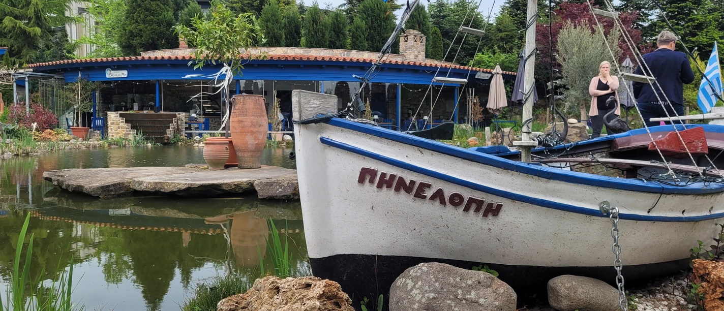 Boot vor einem Teich, umgeben von Pflanzen und einer Terrasse im Hintergrund, mit griechischer Dekoration.