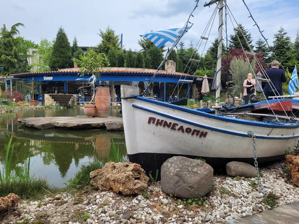 Naturgärten rila erleben Boot vor einem Teich, umgeben von Pflanzen und einer Terrasse im Hintergrund, mit griechischer Dekoration.