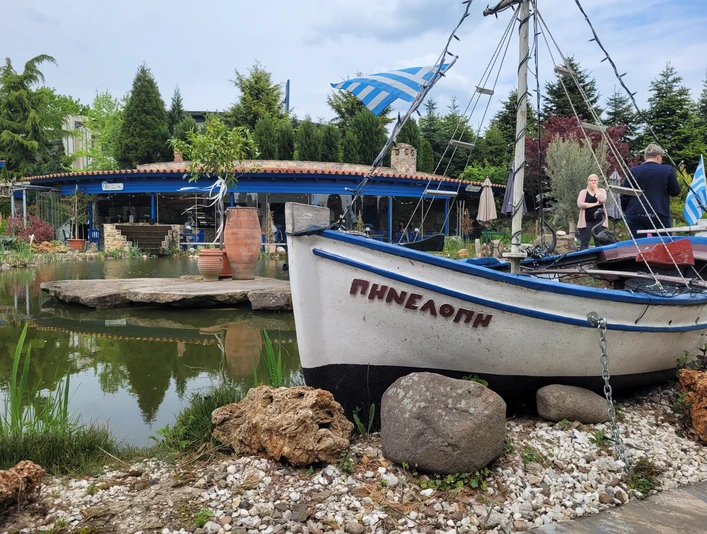 Naturgärten rila erleben Boot vor einem Teich, umgeben von Pflanzen und einer Terrasse im Hintergrund, mit griechischer Dekoration.