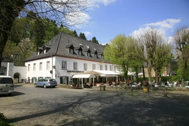Altenberger Hof Historisches Hotel mit Terrasse unter Bäumen, umrundet von Pflastersteinen und Autos vor blauem Himmel.