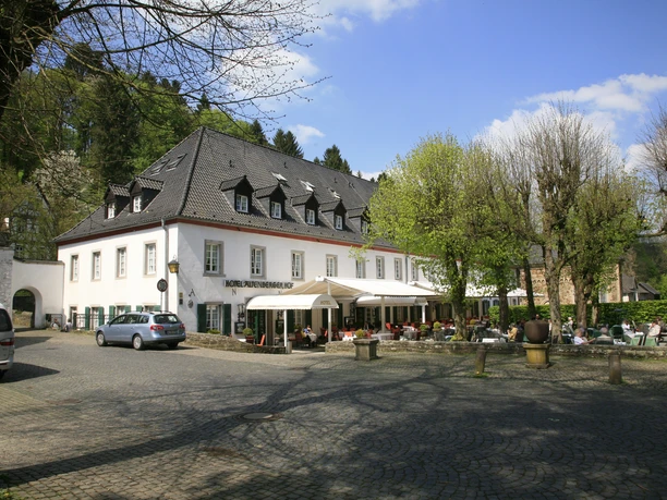 Altenberger Hof Historisches Hotel mit Terrasse unter Bäumen, umrundet von Pflastersteinen und Autos vor blauem Himmel.