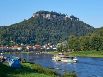 Sächsische Dampfschifffahrt bei Königstein Ein Fluss mit einem Dampfschiff im Vordergrund, dahinter ein bewaldeter Hügel mit der Festung Königstein; Zelte am Ufer, klare, sonnige Atmosphäre.