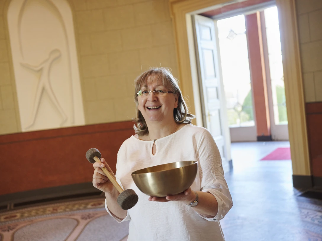 Sigrid Höhr Sigrid Höhr steht in einem hellen Raum mit einer Klangschale und Schlegeln, lächelnd in die Kamera.
