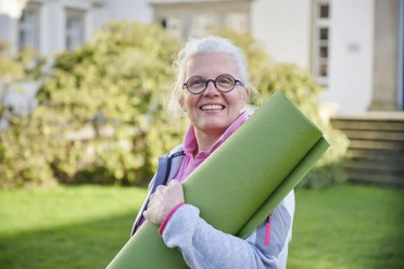 Katja Schwarz Eine Frau mittleren Alters mit weißem Haar und Brille hält eine grüne Yogamatte im Freien.