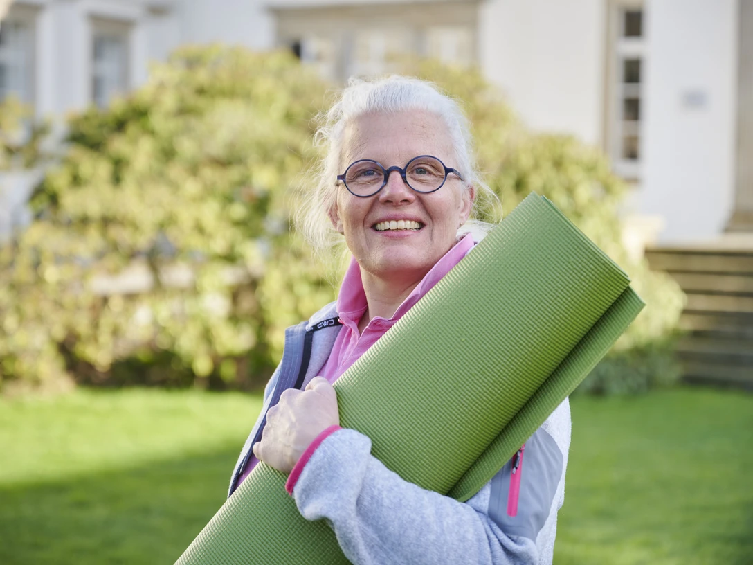 Katja Schwarz Eine Frau mittleren Alters mit weißem Haar und Brille hält eine grüne Yogamatte im Freien.
