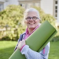Katja Schwarz Eine Frau mittleren Alters mit weißem Haar und Brille hält eine grüne Yogamatte im Freien.