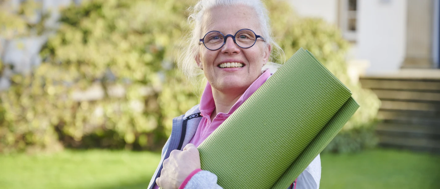 Katja Schwarz Eine Frau mittleren Alters mit weißem Haar und Brille hält eine grüne Yogamatte im Freien.
