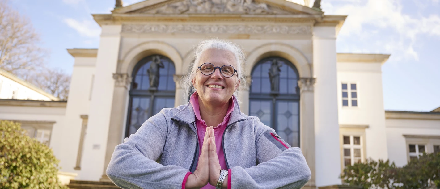 Katja Schwarz Eine lächelnde Frau in Freizeitkleidung macht eine Yoga-Pose vor einem neoklassizistischen Gebäude.