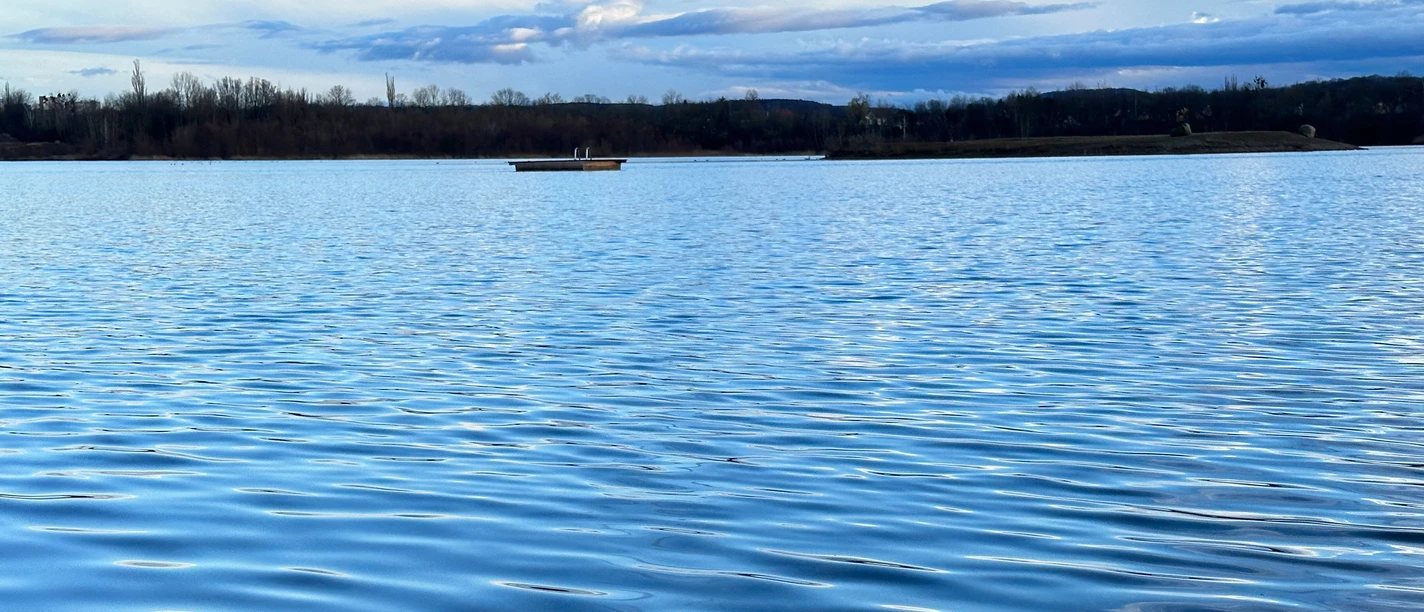 Klidné jezero s modrou vodní hladinou, malým ostrovem v pozadí a zataženou oblohou.