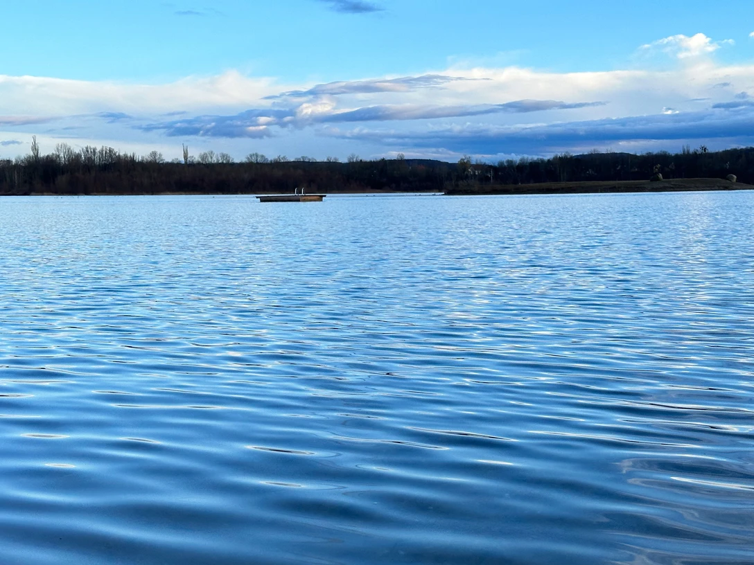 Naturbadesee Pirna-Birkwitz Ruhiger See mit blauer Wasseroberfläche, im Hintergrund eine kleine Insel und bewölkter Himmel.Calm lake with a blue water surface, a small island in the background and a cloudy sky.Klidné jezero s modrou vodní hladinou, malým ostrovem v pozadí a zataženou oblohou.Spokojne jezioro z błękitną taflą wody, małą wyspą w tle i zachmurzonym niebem.Kalm meer met een blauw wateroppervlak, een klein eiland op de achtergrond en een bewolkte lucht.Lago calmo con una superficie d'acqua blu, una piccola isola sullo sfondo e un cielo nuvoloso.