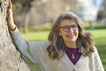Katharina Hinze Eine lächelnde ältere Frau mit grauem Haar und Brille lehnt an einem Baum im sonnigen Park.