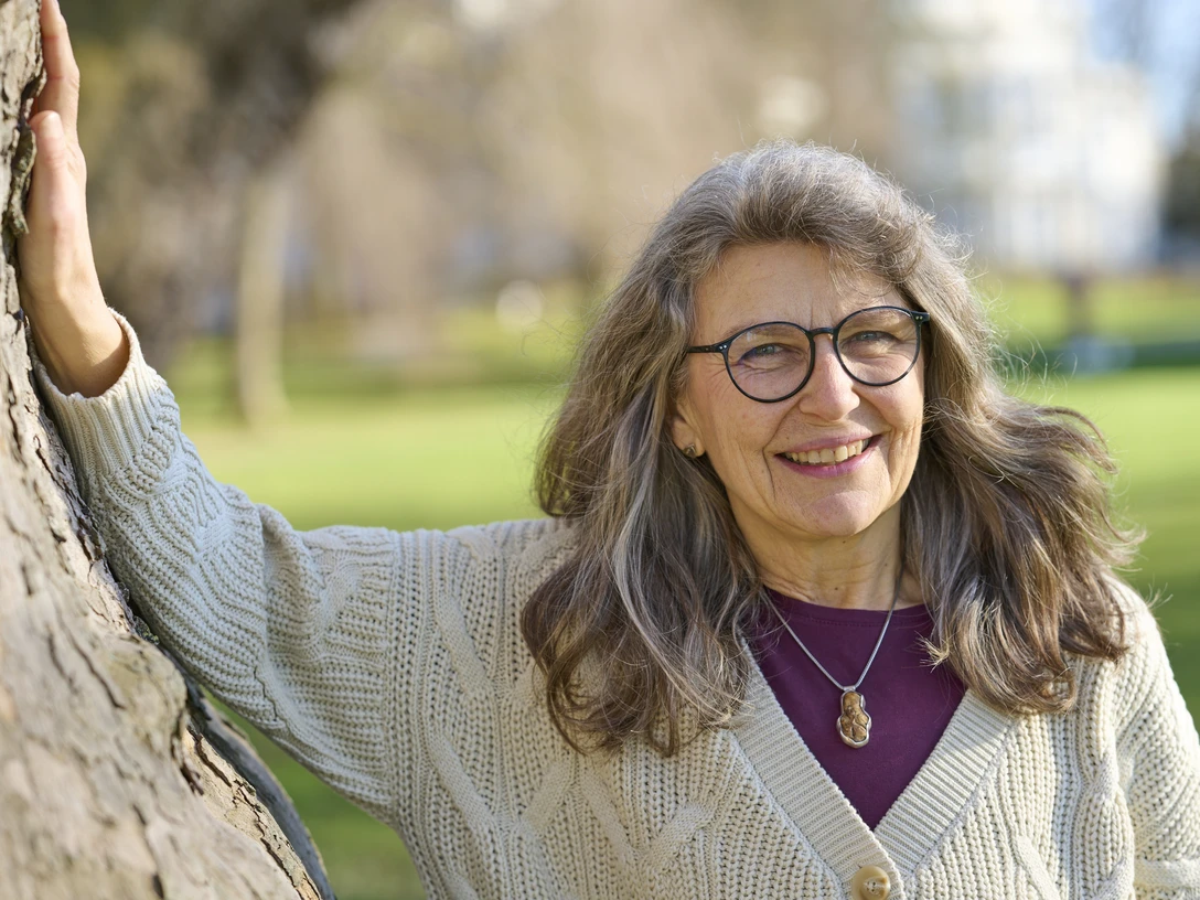 Katharina Hinze Eine lächelnde ältere Frau mit grauem Haar und Brille lehnt an einem Baum im sonnigen Park.